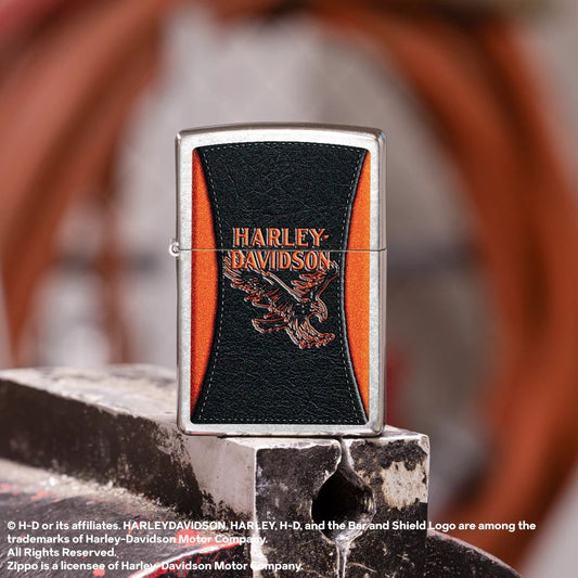 Lifestyle image of Zippo Harley-Davidson® Leathered Eagle Design Street Chrome Windproof Lighter standing on top of a closed workshop vise showing the front of the lighter with a blurred workshop as the background.