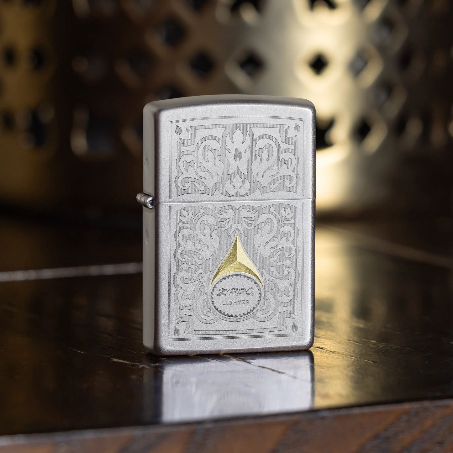 Lifestyle image of Zippo Vintage Filigree Design Satin Chrome Windproof Lighter standing on a dark wooden table showing the front of the lighter with a decorative metal candle holder as the background. 

