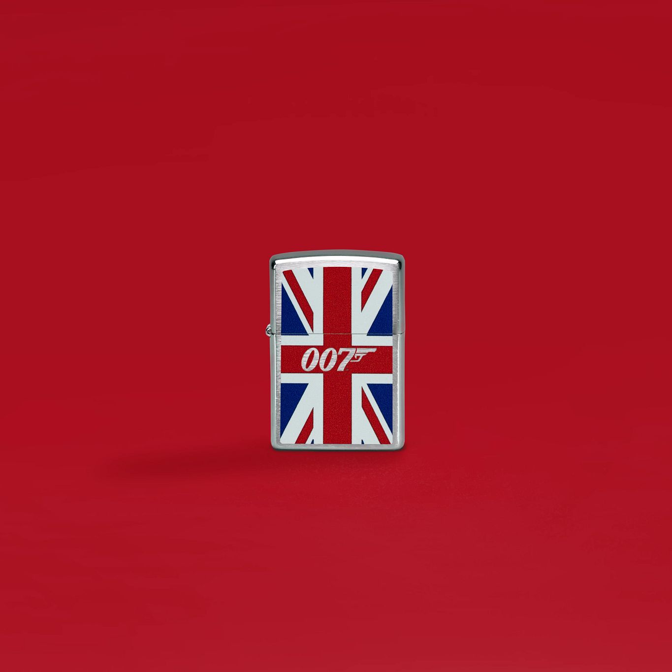Lifestyle image of Zippo James Bond Union Jack Design Brushed Chrome Windproof Lighter standing with a red background.

